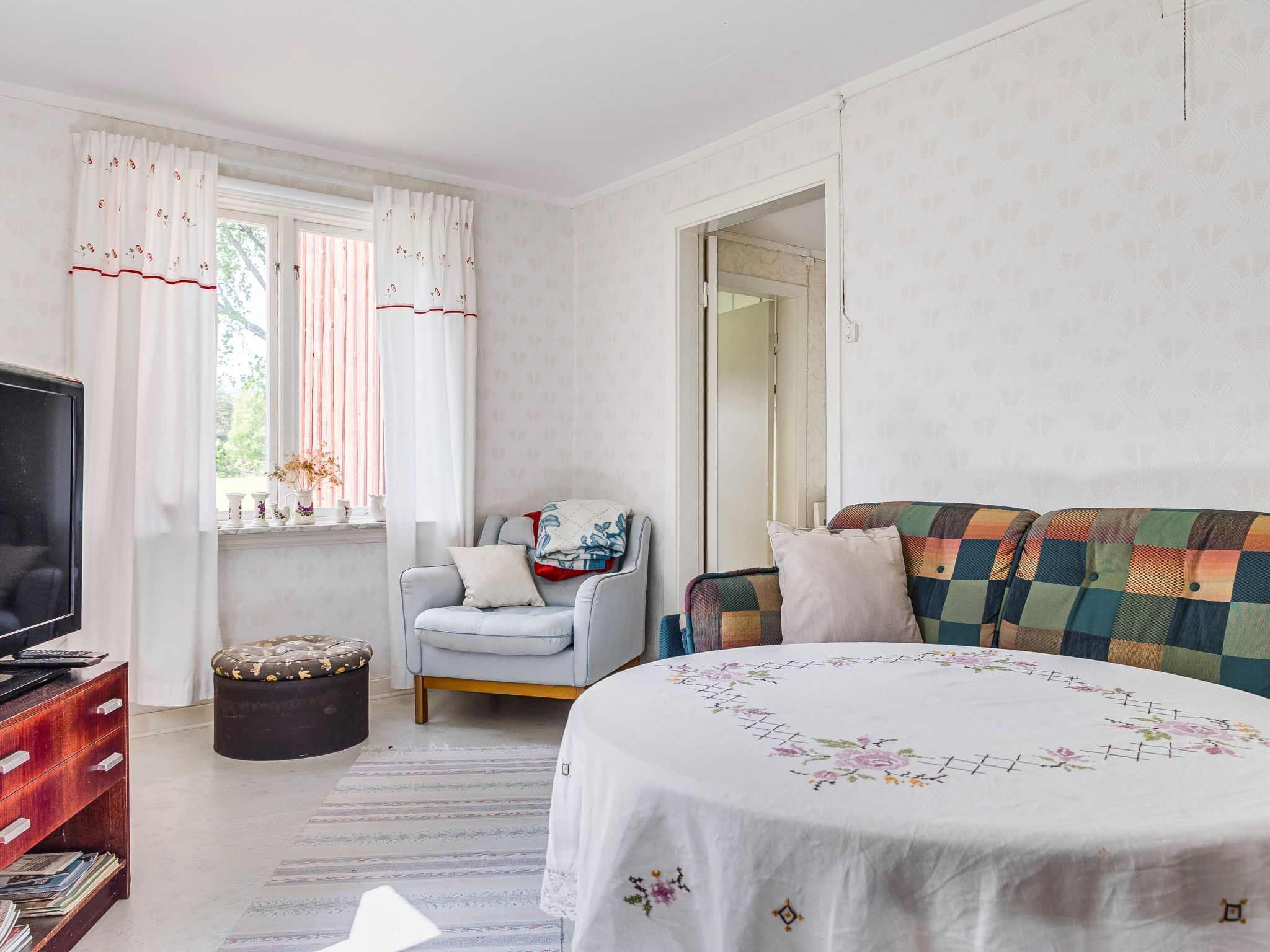 Een slaapkamer met een bed, een stoel, een televisie, een dressoir en een raam met gordijnen.