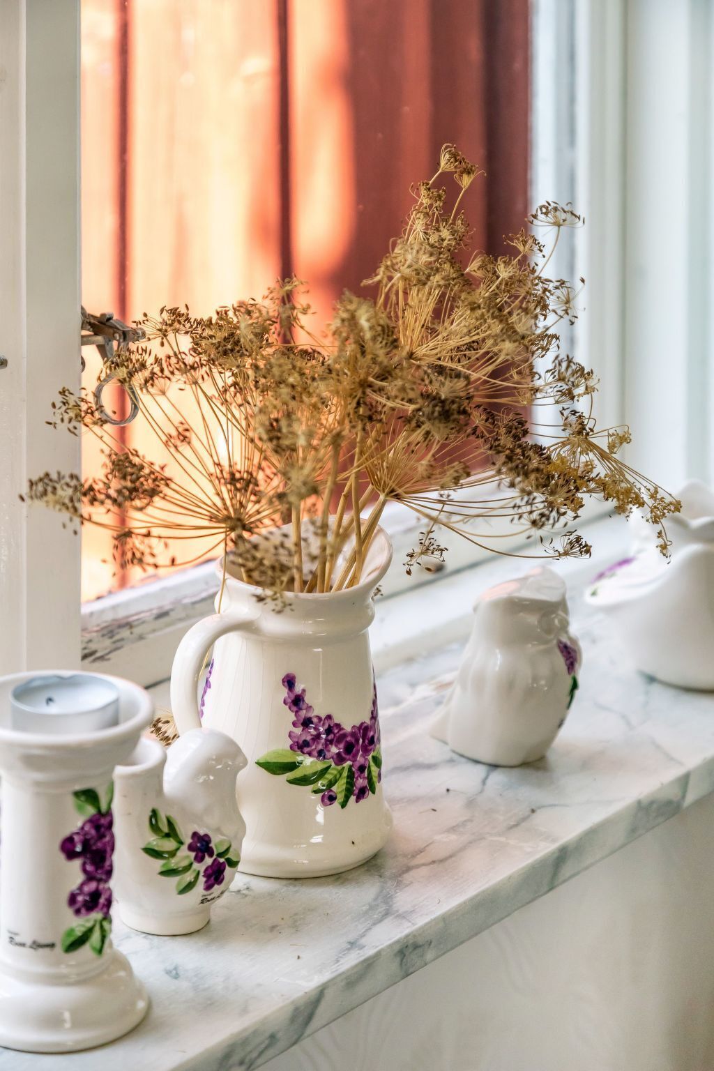 Una jarra de cerámica blanca con flores moradas y plantas secas sobre una encimera de mármol cerca de una ventana.
