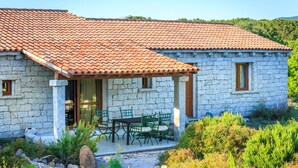A stone house with a tiled roof, a covered patio with a table and chairs, and a garden with various plants. - Mare di Ginepri (BAD115) by Interhome (Badesi)