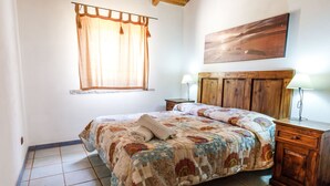 A bedroom with a wooden bed, a window with curtains, a nightstand with a lamp, and a painting on the wall. - Mare di Ginepri (BAD115) by Interhome (Badesi)
