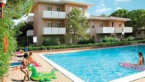 A swimming pool with a clear blue water, surrounded by a brick deck. There are two-story buildings with balconies in the background.