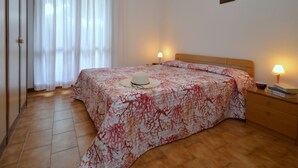 A bedroom with a bed, two bedside lamps, a nightstand, a wooden headboard, a white hat on the bed, and a window with sheer curtains.