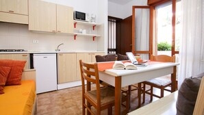 A compact kitchen with wooden cabinets, a microwave, and a small refrigerator. There is a dining table with chairs and a laptop, and a window with curtains.