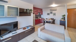 Uma sala moderna com televisão de tela plana, sofá, área de jantar com mesa e cadeiras, cozinha com armários vermelhos e uma porta.