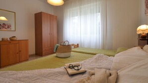 A bedroom with two beds, a wardrobe, a bedside table with a lamp, a window with sheer curtains, and a framed picture on the wall. - Villa Gaby by Interhome (Lignano Sabbiadoro)