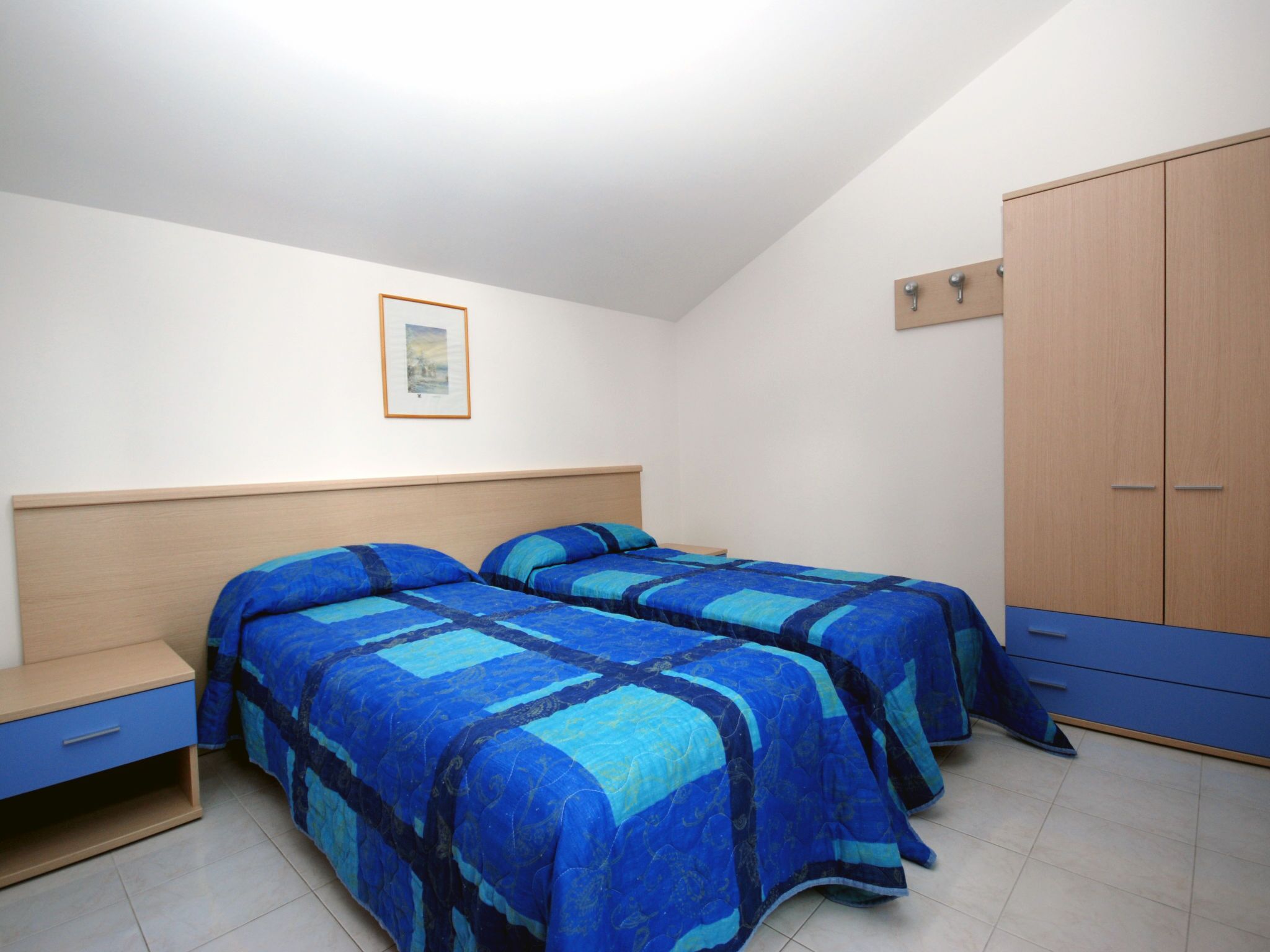 Un dormitori amb dos llits, un armari de fusta, una tauleta de nit i un quadre al mur.