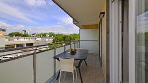 A balcony with a table and chairs, a view of buildings and trees, and a glass door.