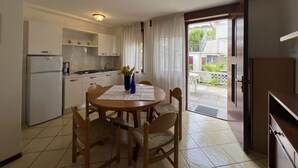 A compact kitchen with white cabinets, a stove, and a refrigerator. There is a dining table with chairs and a vase of flowers. A sliding glass door leads to an outdoor area with a view of buildings and greenery.