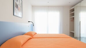 A bedroom with a bed, a nightstand, a wall-mounted lamp, a window with curtains, and a framed picture on the wall.