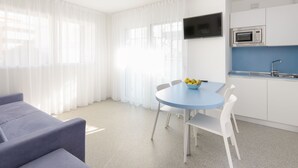 A modern, well-lit room with a round table, white chairs, a sofa, a kitchen area with a microwave, and a TV mounted on the wall. - Residenza Edda by Interhome (Lignano Sabbiadoro)