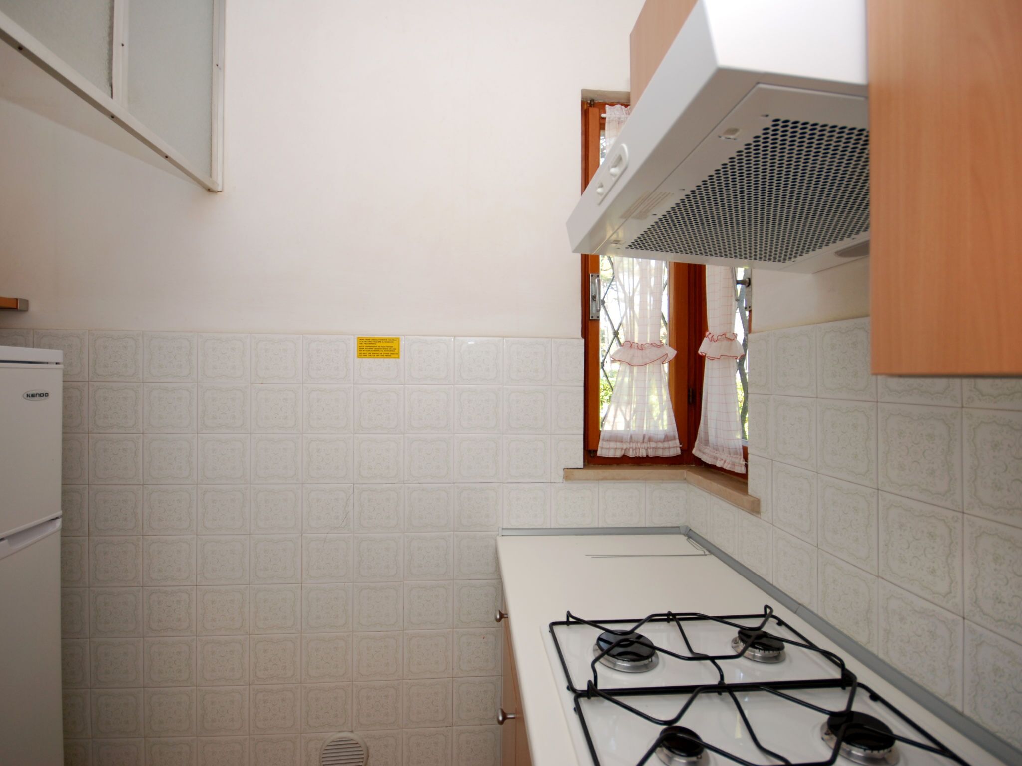Een keuken met een gasfornuis, witte betegelde wanden, een raam met gordijnen en een klein raam met uitzicht.