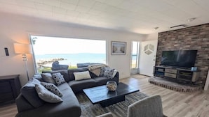 Ett modernt vardagsrum med en stor soffa, ett soffbord och en tv. Genom stora fönster har man utsikt över havet.