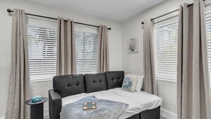 Un dormitorio con una cama grande, dos ventanas con persianas, una mesita auxiliar y una obra de arte colgada en la pared.