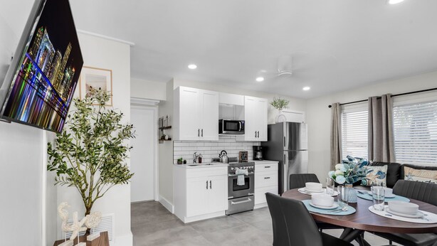 Cocina moderna con armarios blancos, una mesa de comedor lista para cuatro personas y un amplio ventanal con persianas.