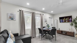 Una sala de estar moderna con sofá, mesa de comedor y sillas. Hay cortinas en las ventanas, un televisor de pantalla plana en la pared y una planta al lado.