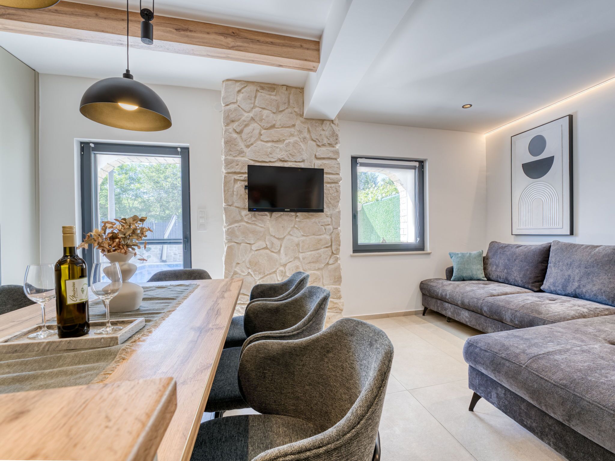 Una sala moderna con una pared de acento de piedra, un televisor de pantalla plana, un sofá gris y una mesa de comedor de madera con sillas.