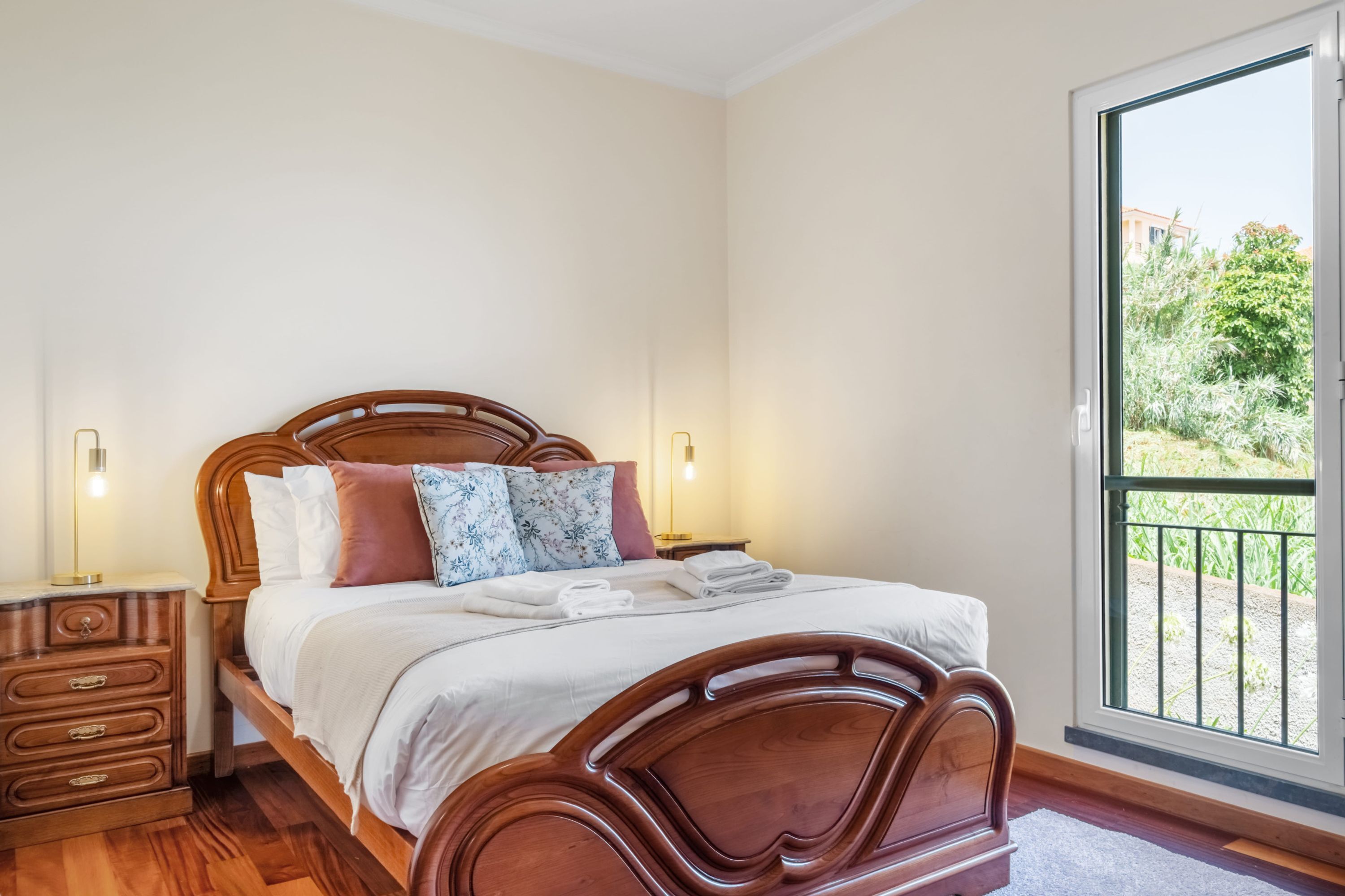 A bedroom with a wooden bed, two bedside tables, a nightstand, and a large window with a view of greenery.