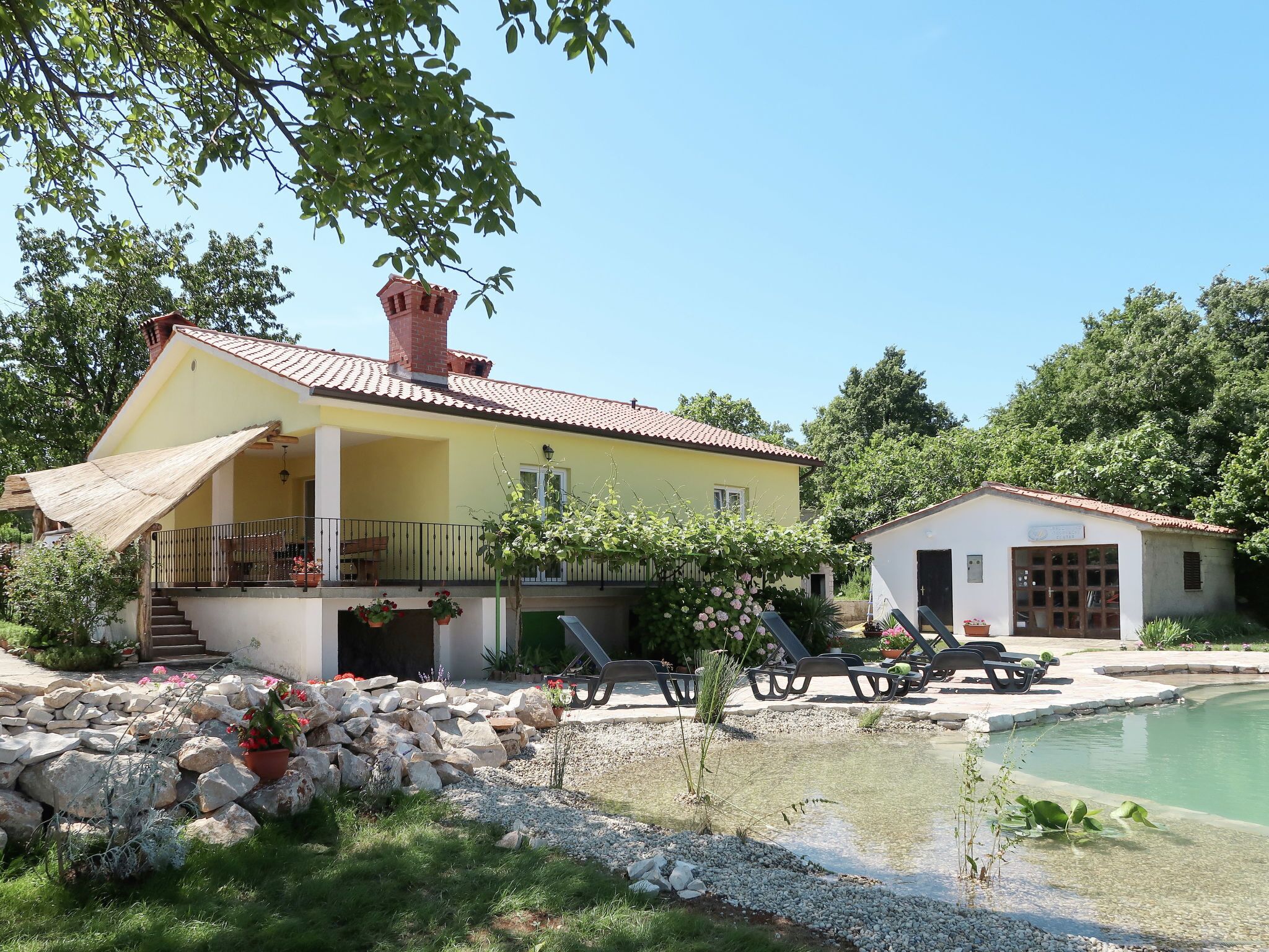 Una casa gialla con tetto rosso, una piccola piscina e un giardino con rocce e piante.