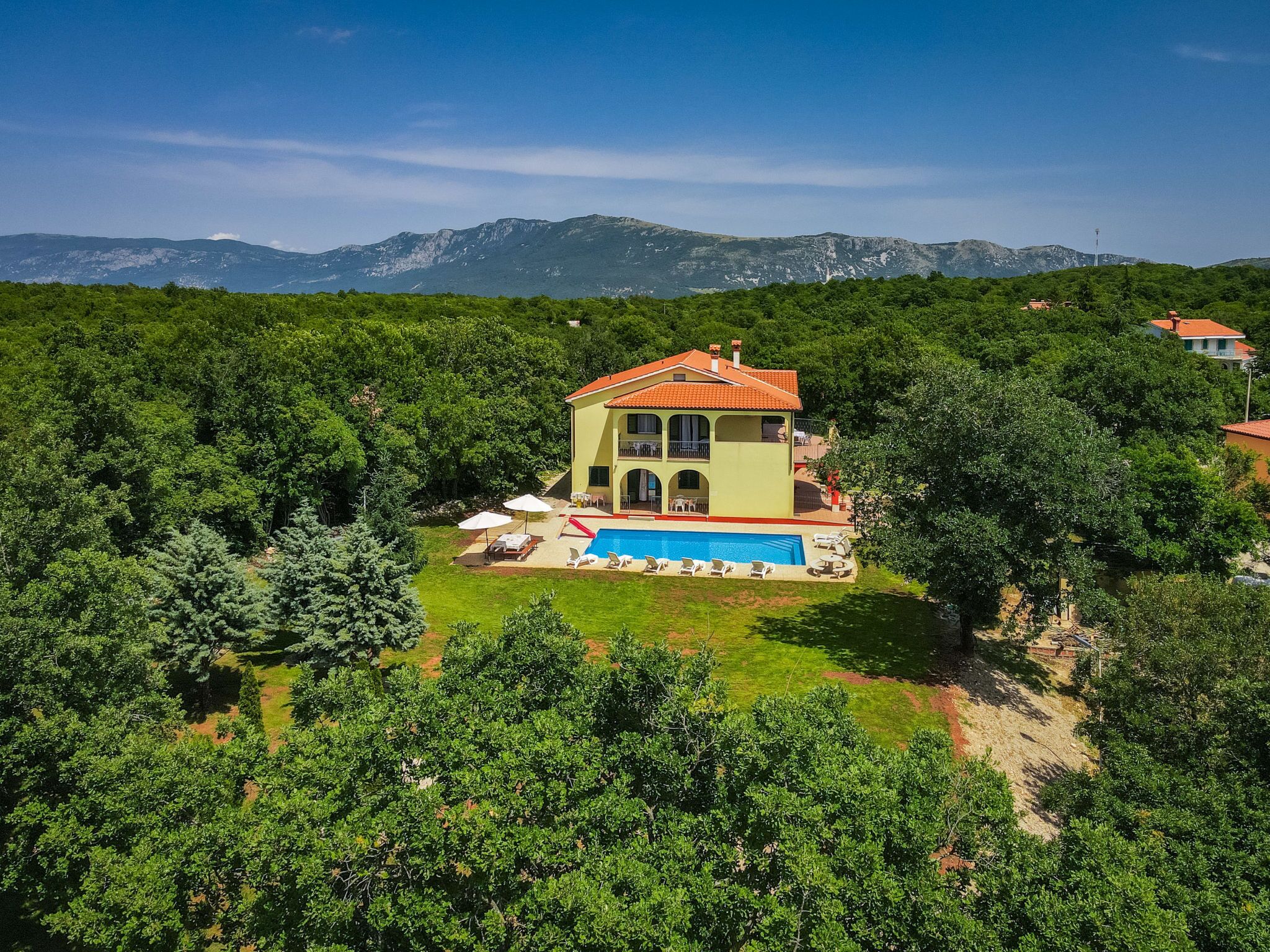A two-story house with a swimming pool, surrounded by dense greenery and mountains in the background.