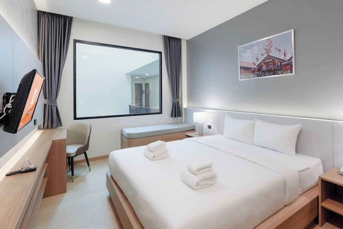 Habitación de hotel con cama, mesitas de noche, escritorio, silla, televisor y un amplio ventanal con cortinas.