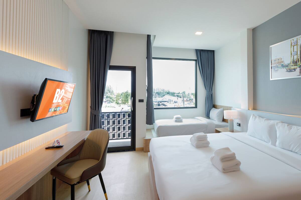 Habitación de hotel con escritorio, dos camas, televisión y un ventanal grande con cortinas.