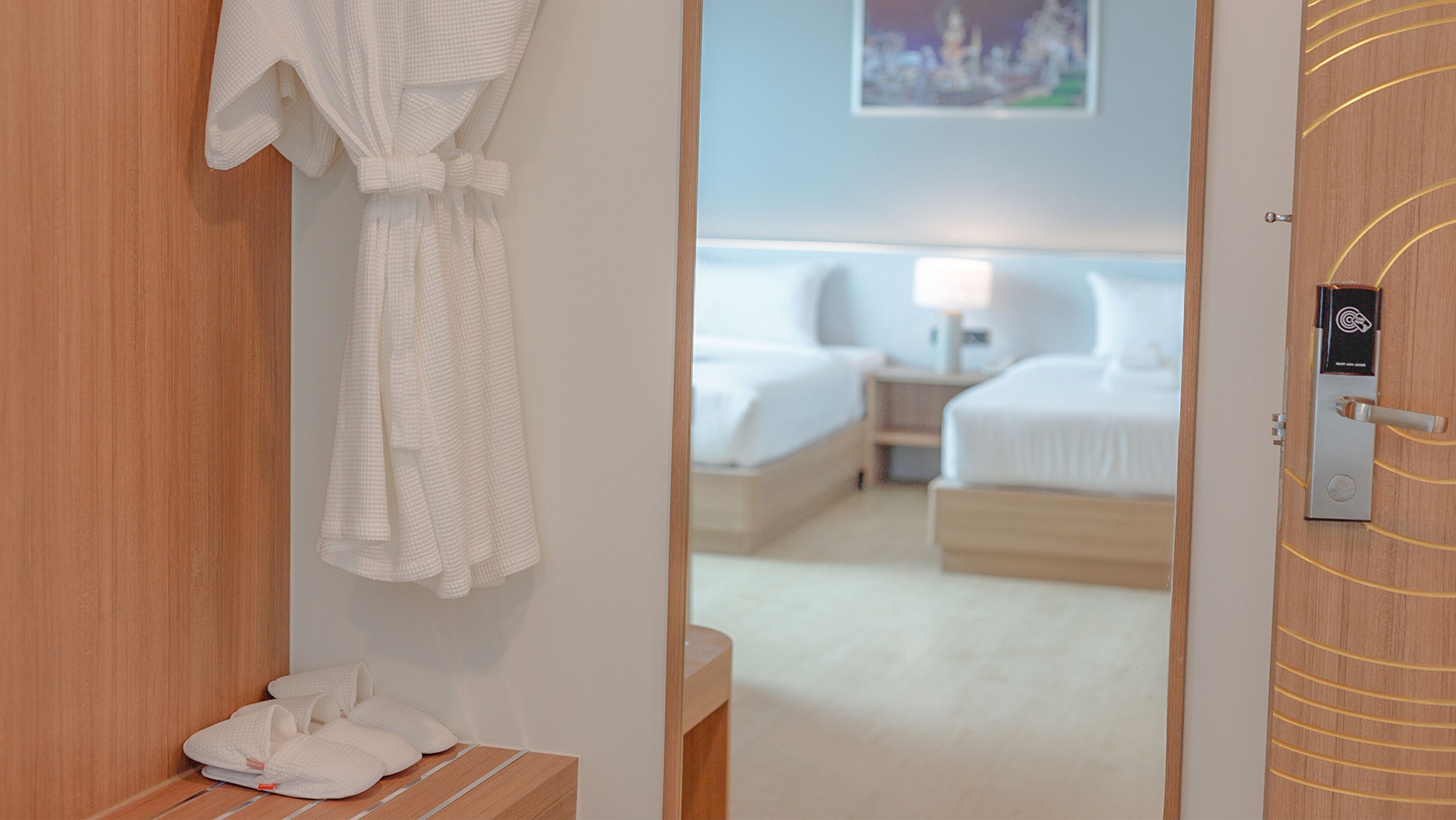 Habitación de hotel con puerta de madera, bata blanca colgada en la pared y cama con ropa blanca.