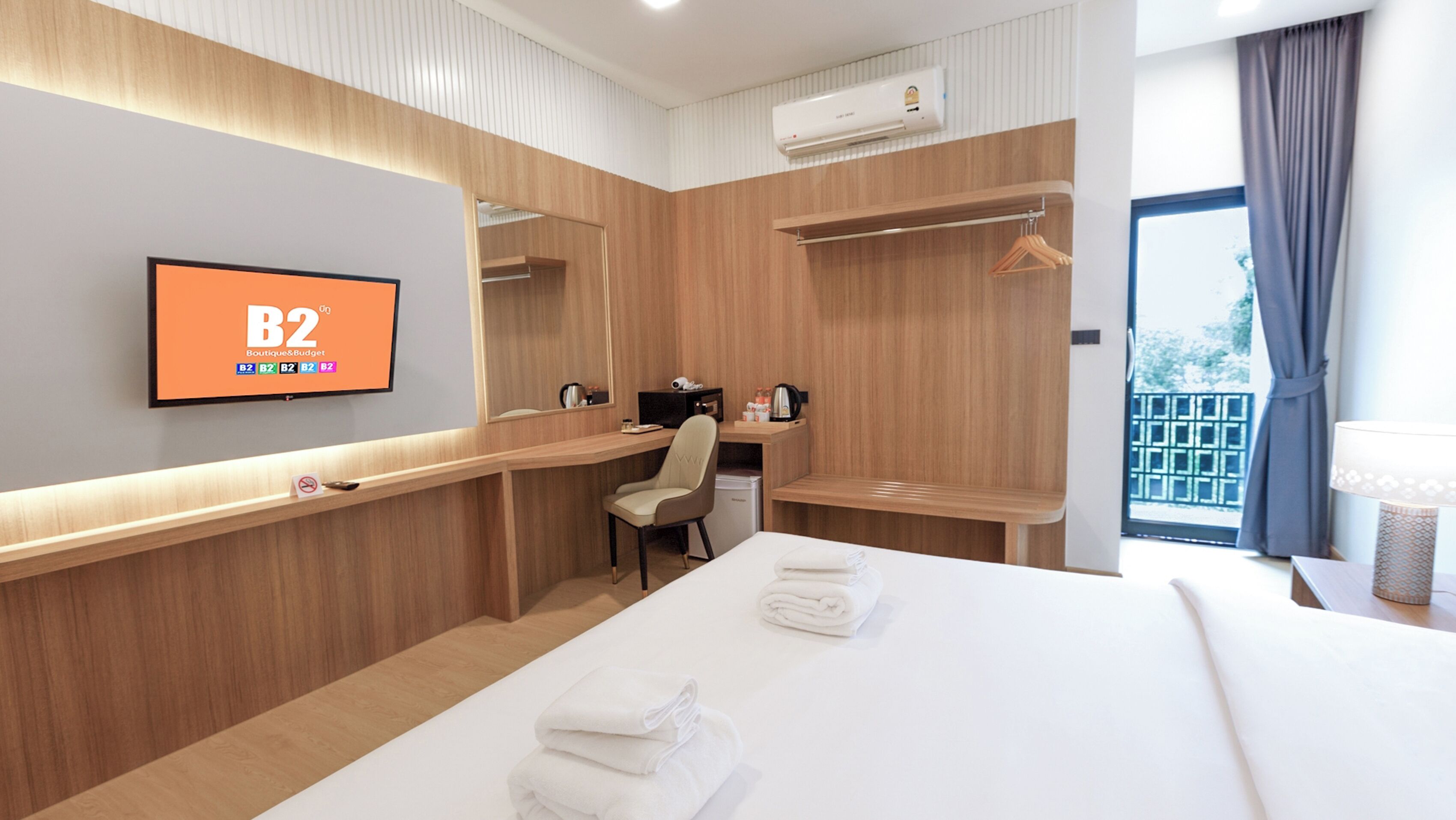 A hotel room with a bed, a desk, a chair, a TV displaying 'B2 Boutique & Budget', and a window with curtains.