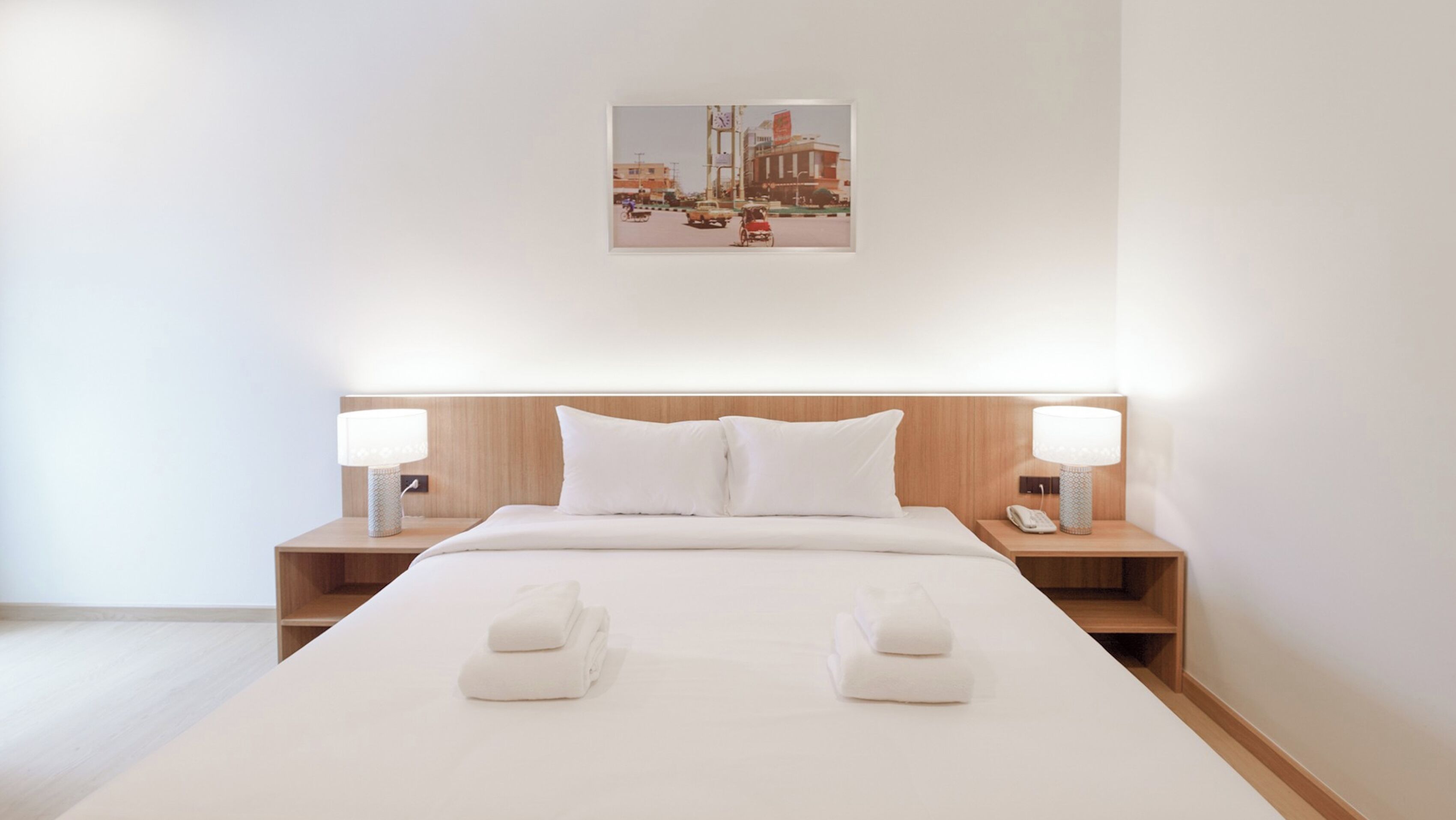 Hotellihuone, jossa on suuri sänky, kaksi yöpöytää valaisimineen, seinälle ripustettu taulu ja kaksi pyyhettä sängyllä.