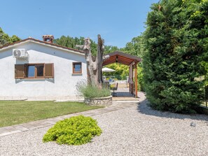 Um edifício branco com pérgola, uma grande árvore perene e um caminho de cascalho.