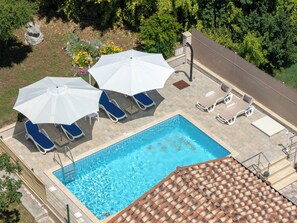 Área de piscina no terraço com espreguiçadeiras azuis e guarda-sóis brancos, cercada por vegetação.