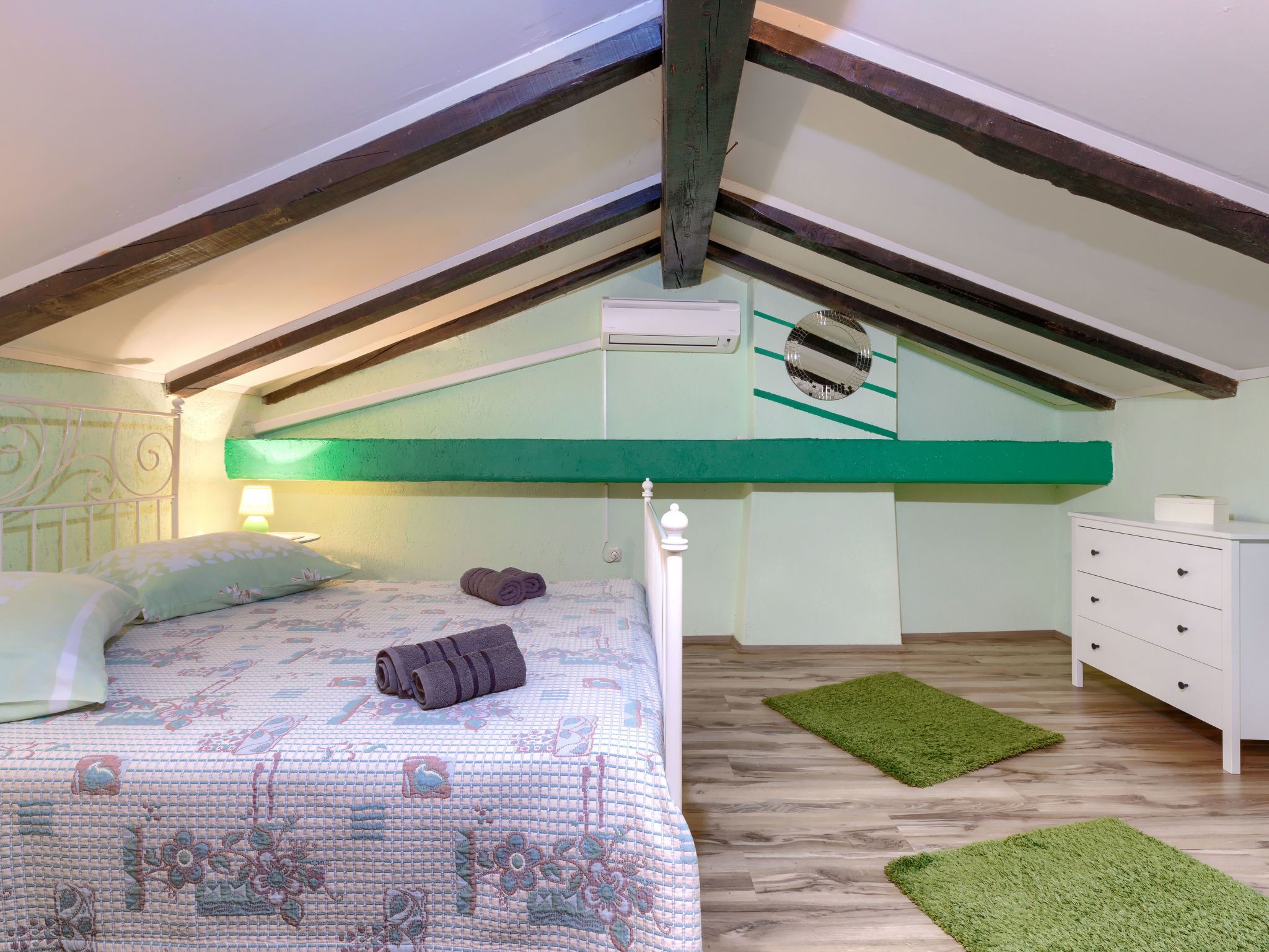 Un dormitorio con techo inclinado, una cama con ropa de cama floral, un tapete verde, un tocador blanco y un equipo de aire acondicionado fijado a la pared.
