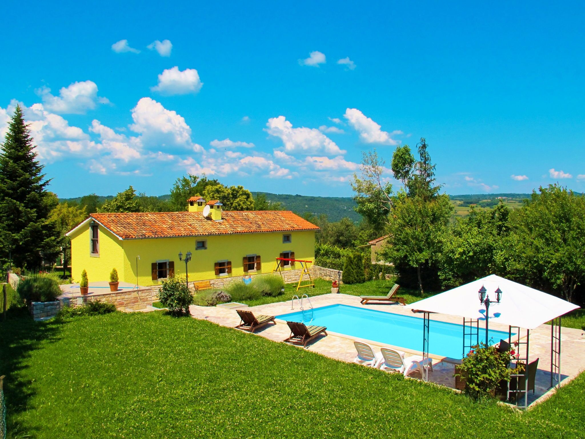 A yellow villa with a swimming pool, surrounded by a lush green lawn and trees.