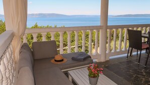 A covered outdoor seating area with a view of the sea, a white balustrade, and a potted plant.