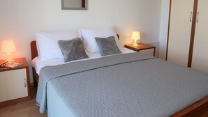 A neatly made bed with a grey quilt, two bedside lamps, a nightstand, a wooden wardrobe, and a mirror on the wall.