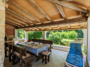 A covered outdoor dining area with a table and benches, a brick oven, and a view of greenery.
