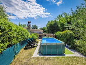 Une piscine entourée de chaises longues, bordée de verdure, et une maison avec une cheminée en pierre.