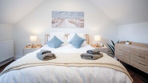 A bedroom with a high ceiling, a large bed with two pillows, a bedside table with a lamp, and a wooden dresser. - The Carthouse - 1 Bedroom Cottage - Princes Gate (Narberth)