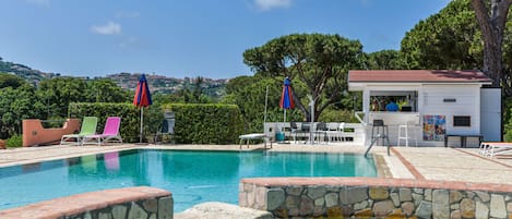 Ein Schwimmbecken mit steinernen Bordüren, Liegestühlen und Sonnenschirmen.