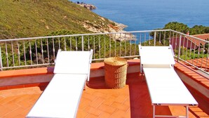 A terrace with two white lounge chairs, a wicker basket, and a view of the sea and a rocky coastline. - Panorama by Interhome (Capoliveri)