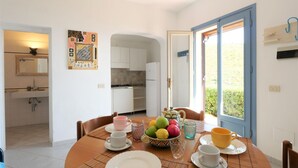 Un coin repas avec une table ronde dressée pour quatre personnes, une cuisine en arrière-plan et une porte donnant sur l’extérieur.