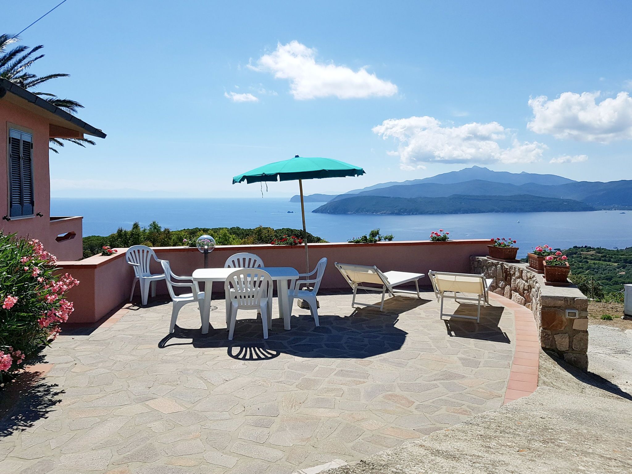 Eine Terrasse mit Tisch und Stühlen, einem grünen Sonnenschirm und Blick auf Meer und Berge.