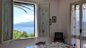 A room with a table covered in a patterned tablecloth, a television on a small table, and a view of the sea through a large window. - Apartment Palma by Interhome (Capoliveri)