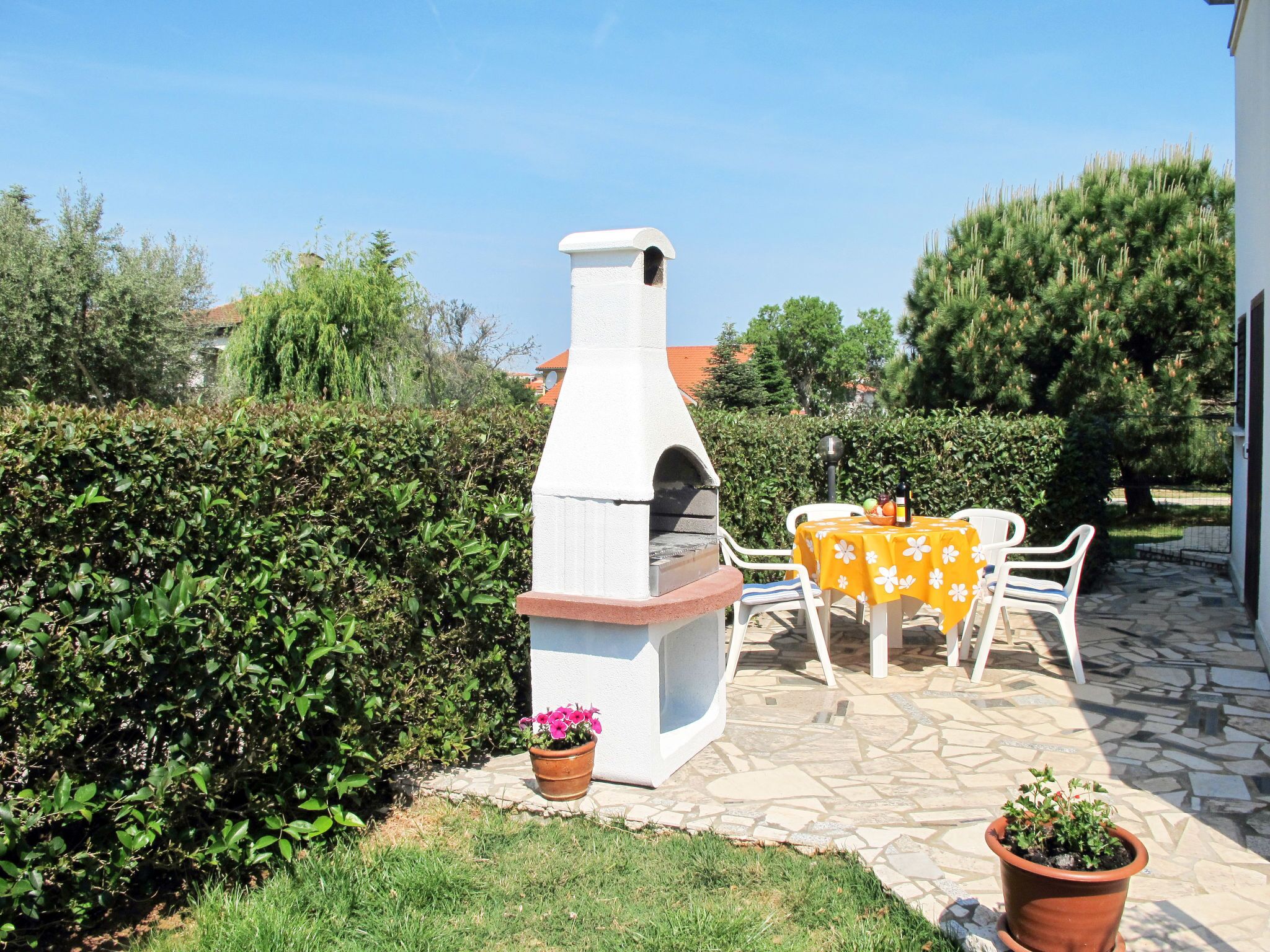 An outdoor patio with a stone-tiled floor, a white barbecue oven, a table with a yellow tablecloth, and potted plants.