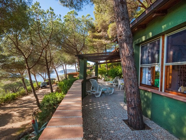 A covered outdoor seating area with chairs and a table, surrounded by trees and a view of the sea.