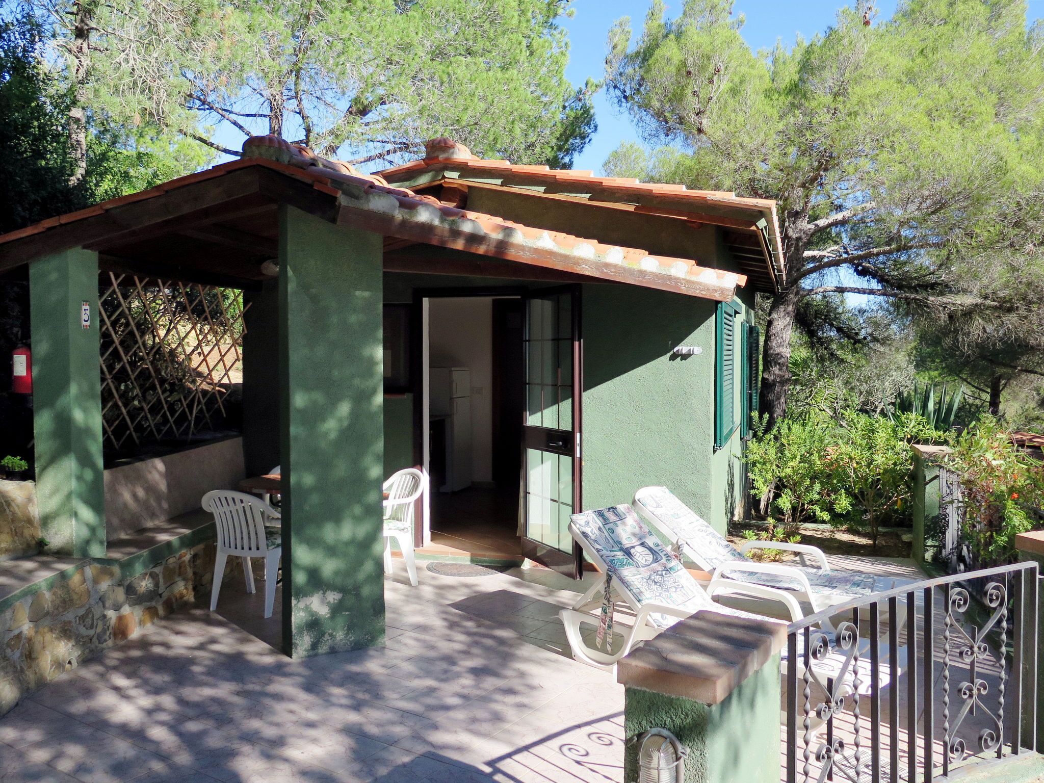 A small, green-roofed building with a patio area, outdoor seating, and a view of trees.