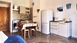 A compact kitchen with a dining table, chairs, a refrigerator, a coffee maker, and a stove. - BINI (REI210) by Interhome (Costa Rei)