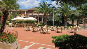 An outdoor patio area with tables and chairs, surrounded by palm trees and a building with a tiled roof. - Mirto Marino (REI410) by Interhome (Costa Rei)