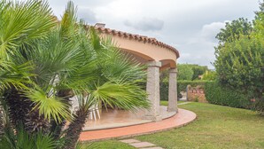 A covered outdoor area with a curved stone pathway leading to a seating area, surrounded by lush greenery and a stone wall. - Rei Sole (REI303) by Interhome (Costa Rei)