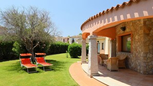 A villa with a stone facade, a covered patio with wicker furniture, and a well-maintained lawn with two red lounge chairs. - Rei Sole (REI301) by Interhome (Costa Rei)