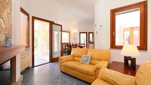 A living room with a yellow sofa, a dining area with chairs, and a kitchen area with a stove. - Rei Sole (REI301) by Interhome (Costa Rei)